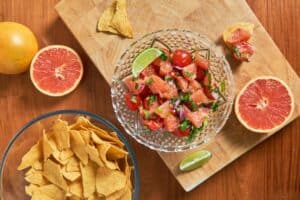 A glass bowl of grapefruit salsa with cherry tomatoes, onions, and cilantro on a wooden cutting board, alongside a bowl of tortilla chips and halved grapefruits, with a wooden tabletop background.