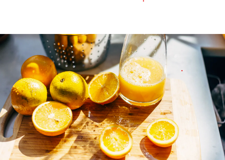 Freshly squeezed orange juice in a glass, with whole and halved oranges on a wooden cutting board, and a metallic juicer in the background, all under bright lighting.