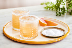 Two grapefruit Paloma cocktails with salted rims on a round yellow tray, accompanied by a small plate with salt and two grapefruit wedges on the side.